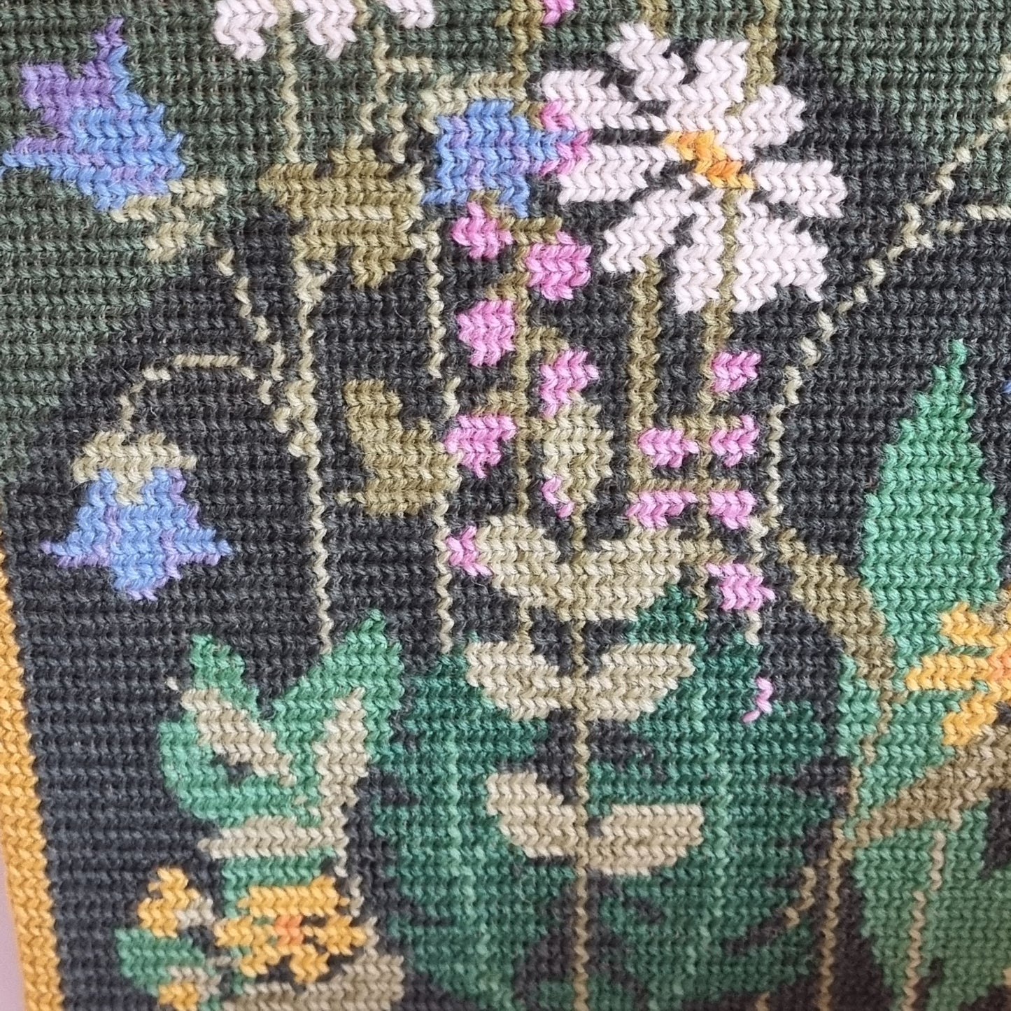 Close-up detail of lilac, blue and white flowers in needlepoint on forest green wool canvas