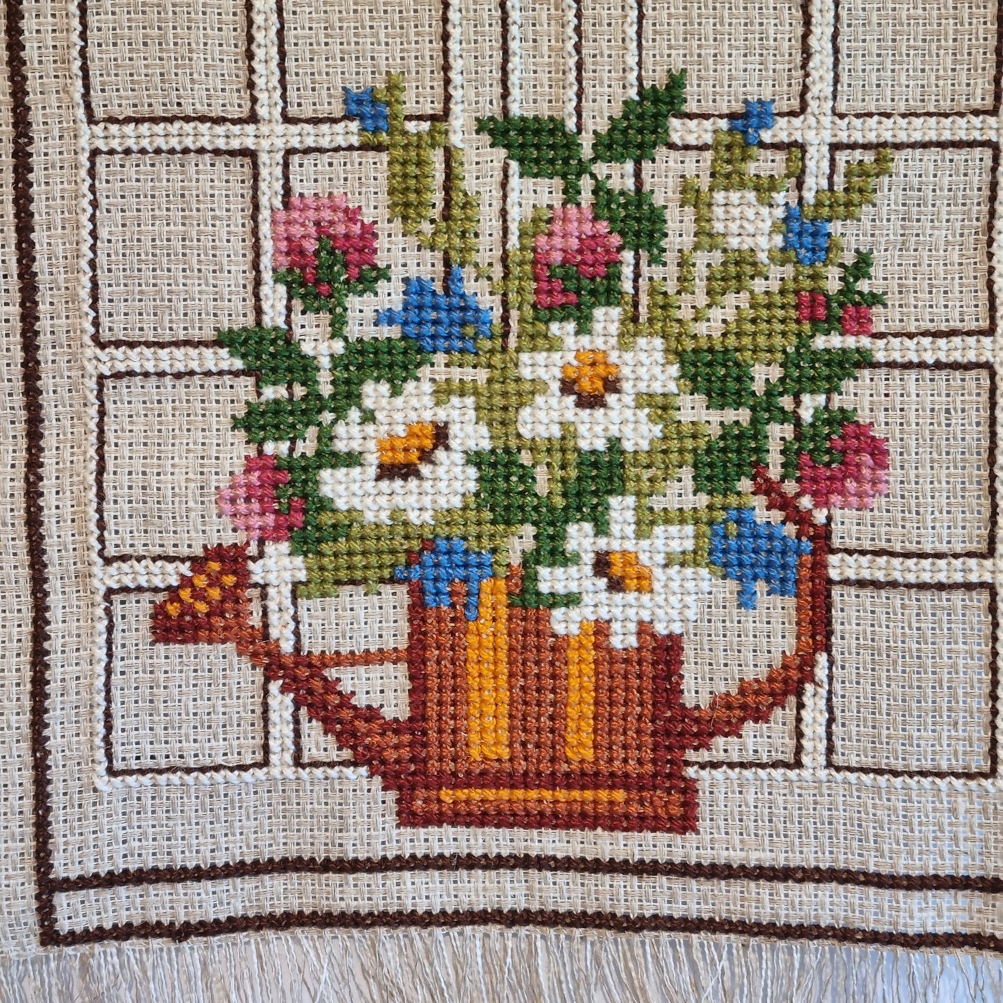 Close-up detail of cross-stitch flower bouquet in watering can, pink roses, blue cornflowers and white daisies