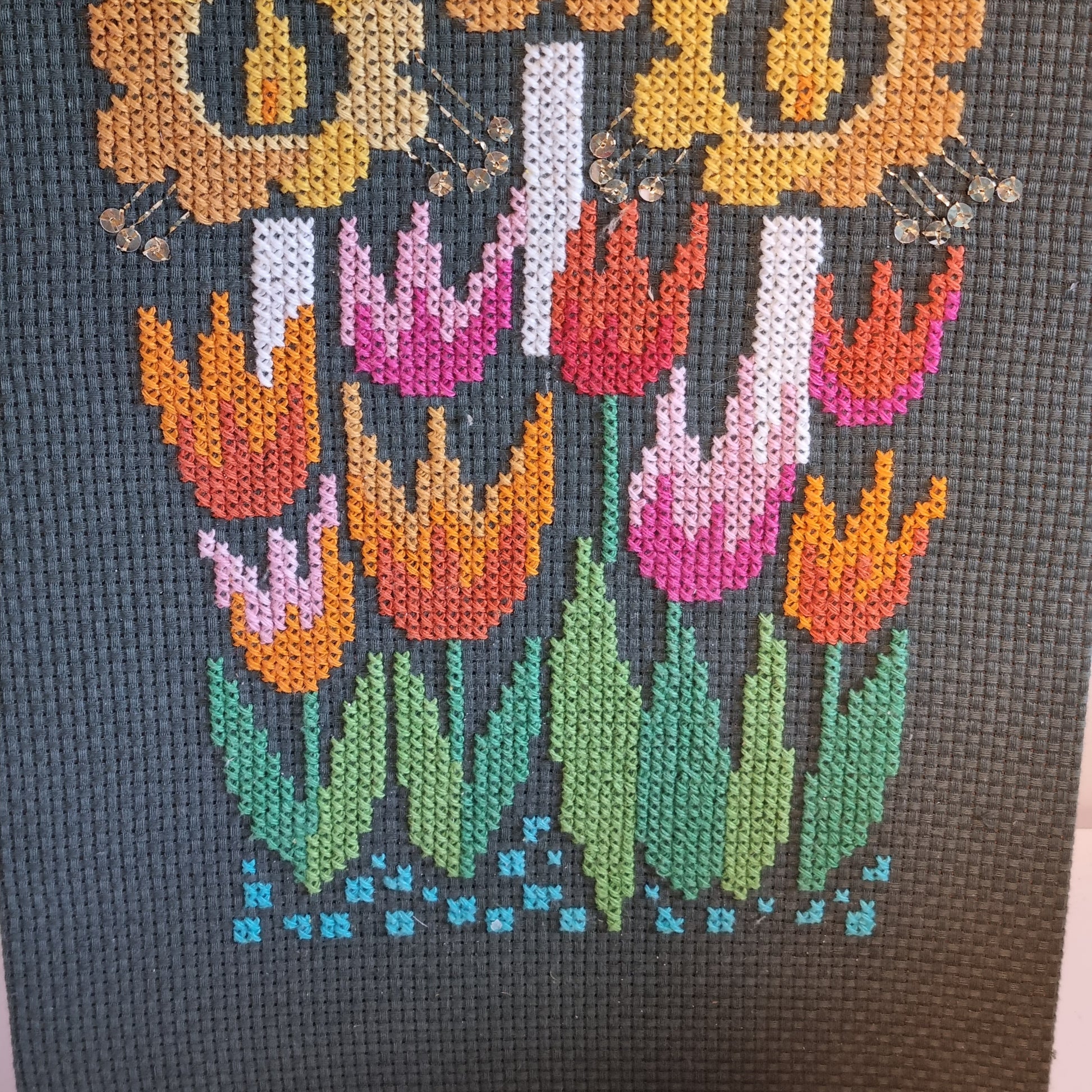 Close-up detail of cross-stitch tulips in orange, pink and red on charcoal grey canvas with turquoise border