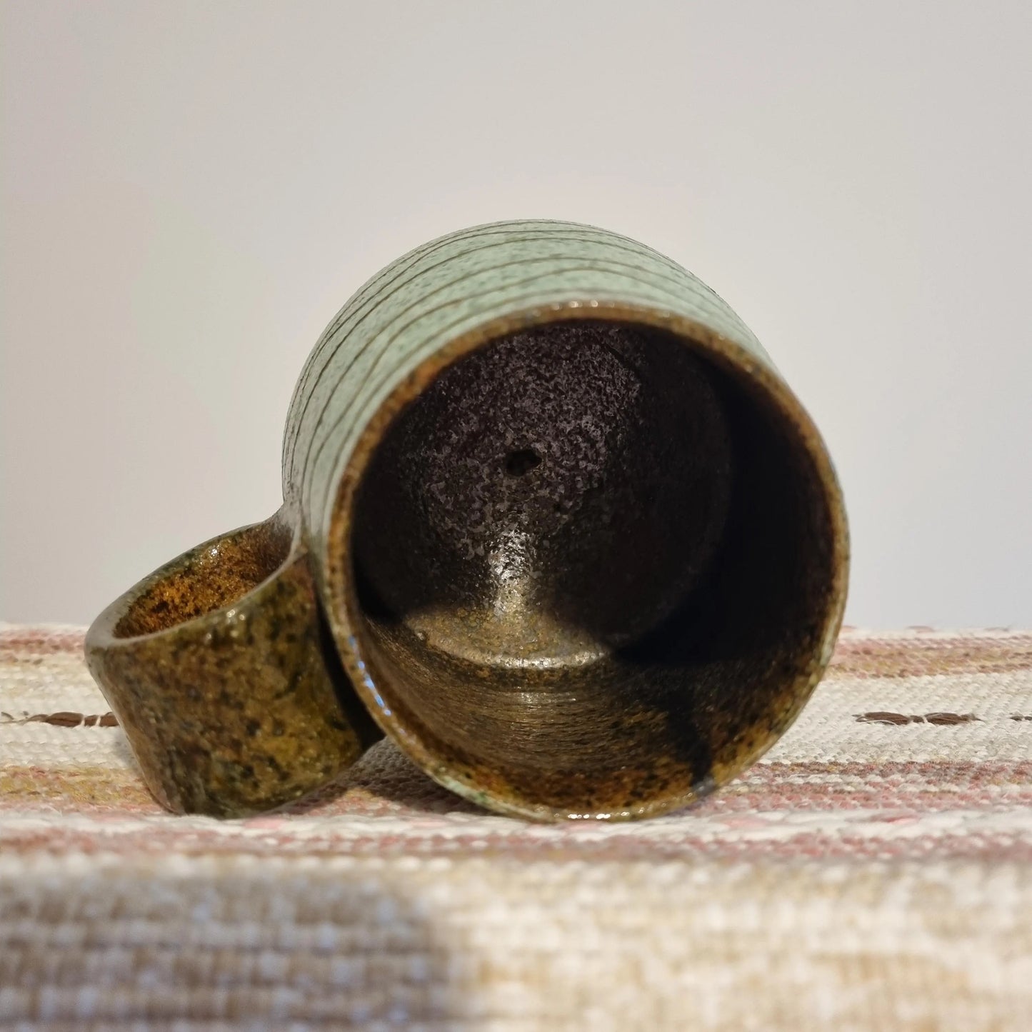 Krukmakeri Ystad Swedish Stoneware Mug – Speckled Green Glaze, Handmade, lying on its side on a woven fabric surface