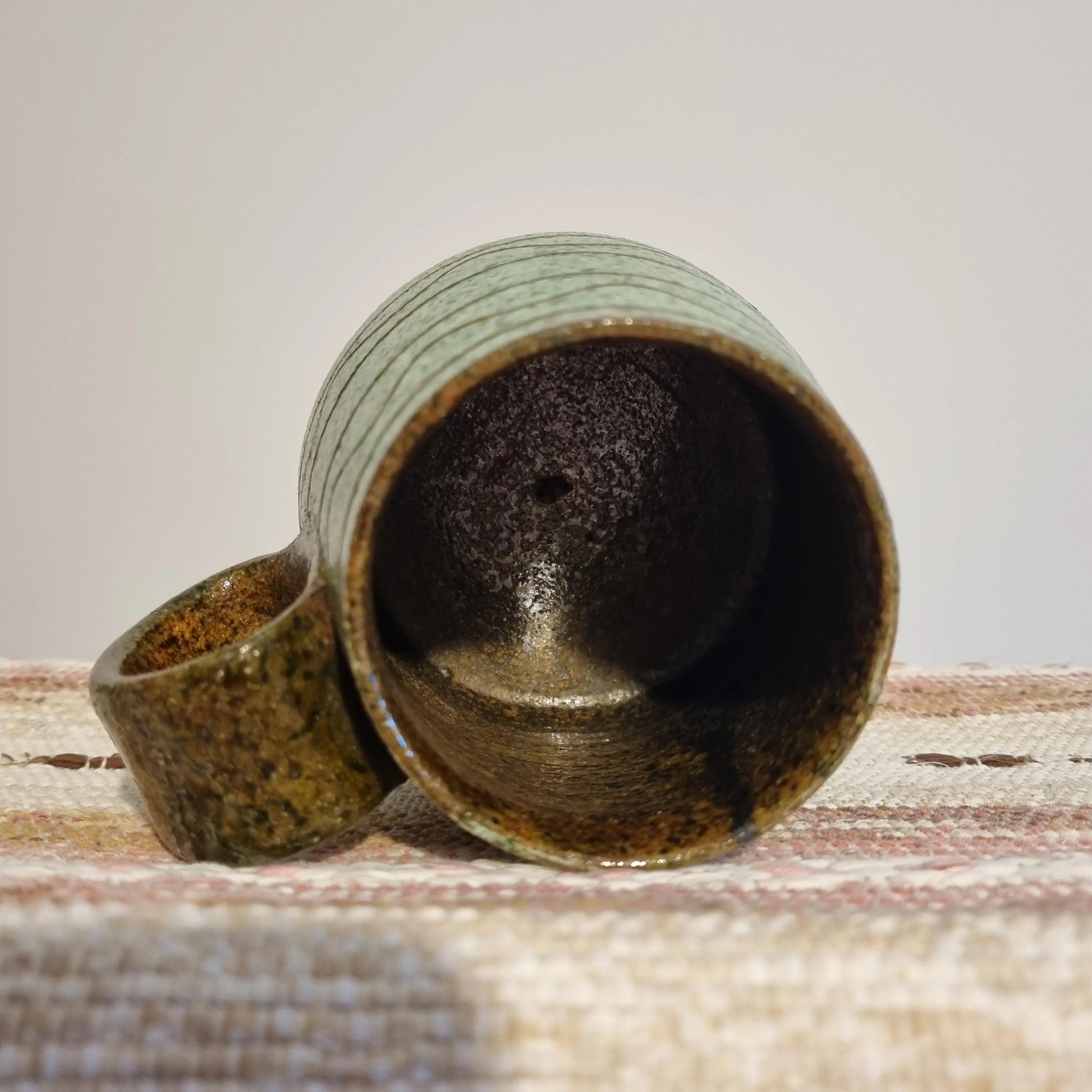 Krukmakeri Ystad Swedish Stoneware Mug – Speckled Green Glaze, Handmade, lying on its side on a woven fabric surface