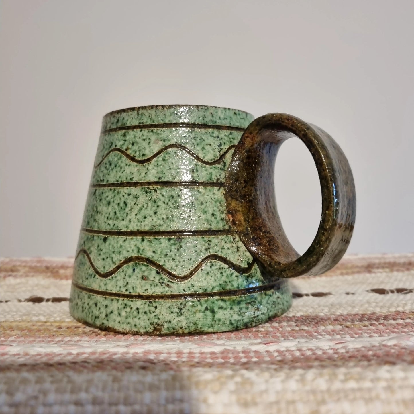 Krukmakeri Ystad Swedish Stoneware Mug – Speckled Green Glaze, Handmade with wavy line patterns and a rounded handle on woven cloth.
