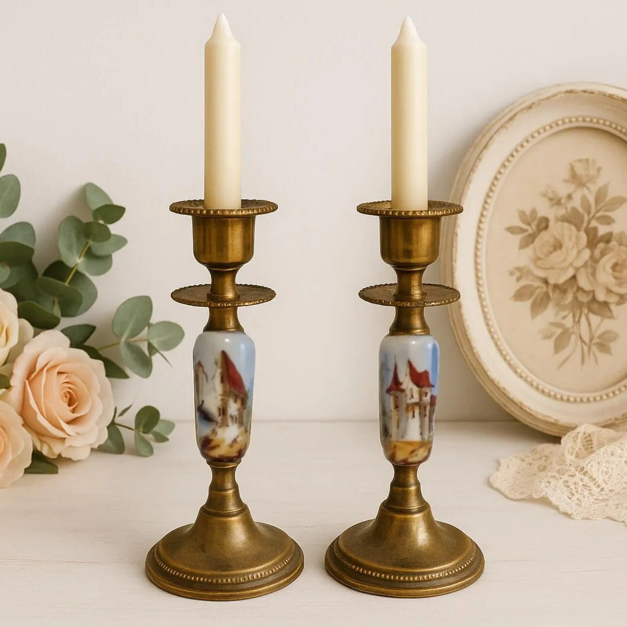 Vintage Brass Candle Holders: Hand-Painted European Village Scene, 1920s Pair with white candles and floral decor background.